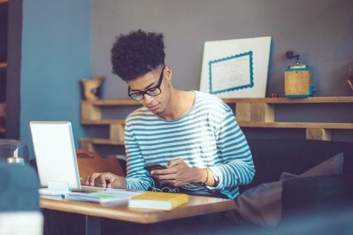 Young man sits at home checking his mobile phone as he works on his laptop