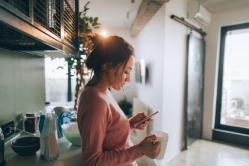 Woman looks at phone while drinking coffee in her kitchen.