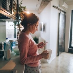 Woman looks at phone while drinking coffee in her kitchen.