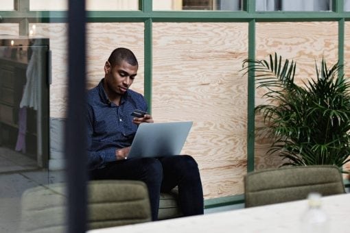 Young man looks thoughtfully at his credit card as he sits with a laptop on his legs