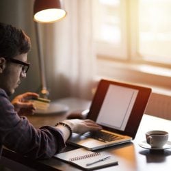 Man working at desk on laptop with a notepad, pen, and coffee mug nearby.