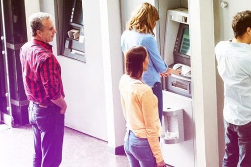 Five people lined up to use four ATMs on the street. Three people are currently using the ATMs. A man and a woman are waiting for their turns.