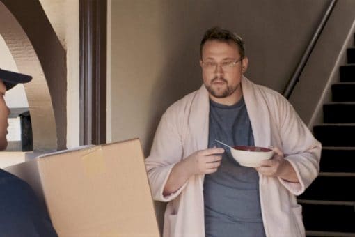 Man in a bathrobe and eating cereal gets a package from a delivery man.