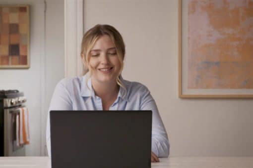 A woman sits and smiles as she looks down at her laptop screen.