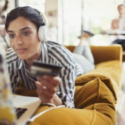 Young woman lays on her couch holding credit card and looking at laptop.