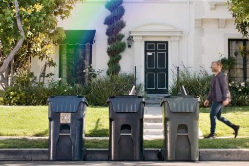 Three trash cans are lined up, with a rainbow emerging from one, as a man approaches.