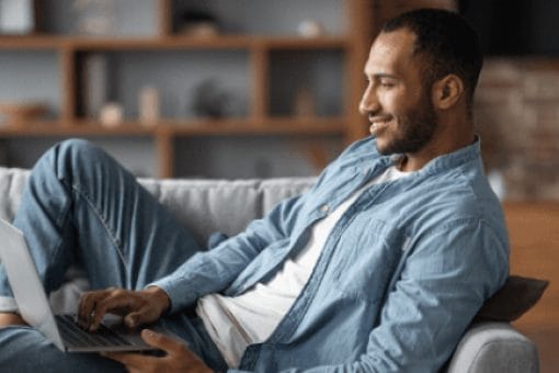 Man in blue shirt smiles looking at laptop as he reclines on a couch