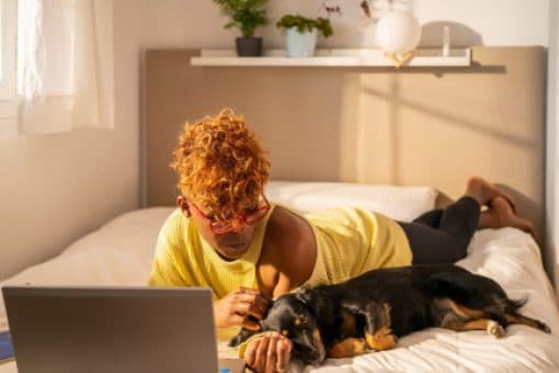 A woman petting her dog on a bed while using a laptop.