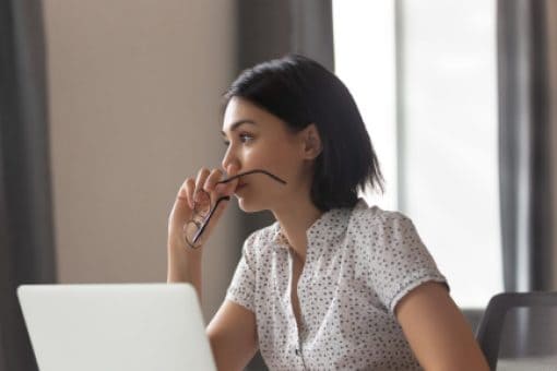 Asian woman with a contemplative expression sitting in front of a laptop and staring into the distance.