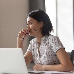 A woman with a contemplative expression sits in front of a laptop and stares into the distance.