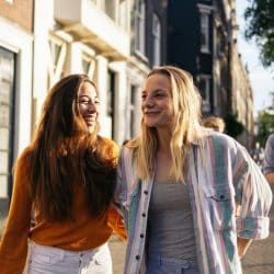 Two young women laugh and talk while walking down a busy sidewalk in a shopping district.