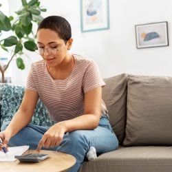 A young woman sits on her sofa and works on calculating her income with pen and paper.