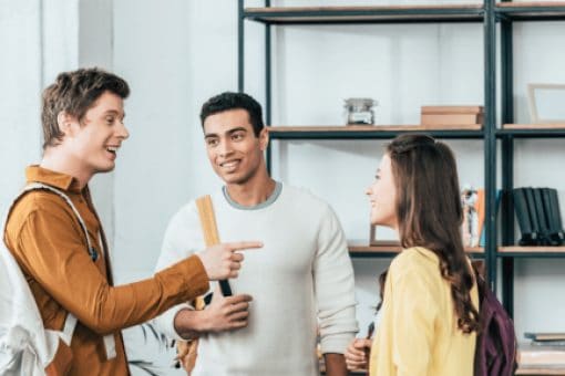 Three students with backpacks have an animated conversation in front of a bookshelf.