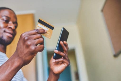 A man looks down at a credit card held in one hand and mobile phone in the other hand.