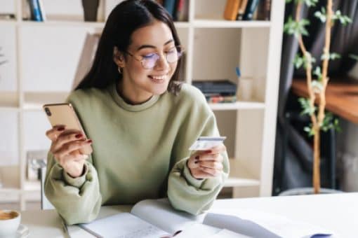 A young college student smiles at a credit card in one hand while holding a mobile phone in the other