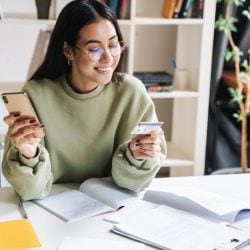 A young college student smiles at a credit card in one hand while holding a mobile phone in the other