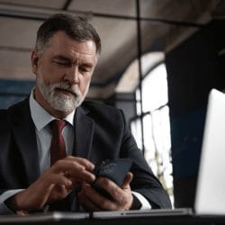 A man in a suit and tie uses his mobile phone and laptop in his office.