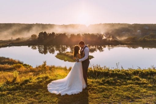 Bride and groom stand near a lake at sunset