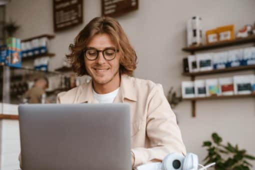 Young long-haired smiling man in glasses working with laptop while sitting with coffee in cafe