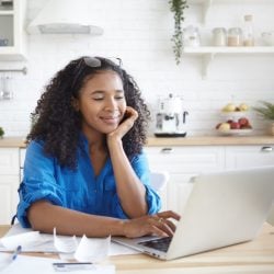 Smiling woman uses laptop at kitchen table covered with papers, a pen, and a coffee cup.