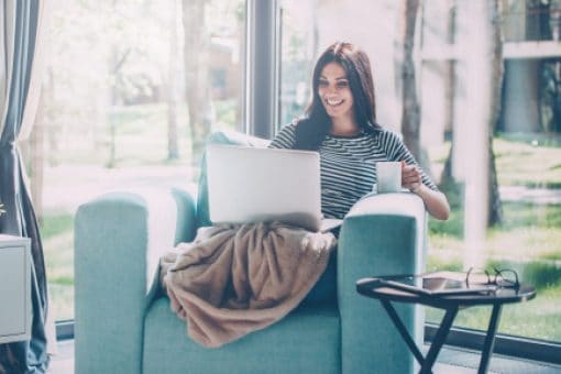 Smiling young woman, her legs covered with a blanket, sits in a chair looking at her laptop and holding a cup of coffee.