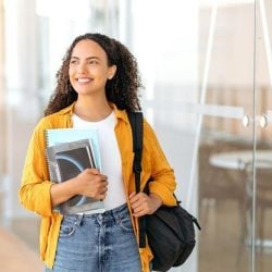 A student carrying notebooks and wearing a yellow shirt stands in a modern building with glass doors.