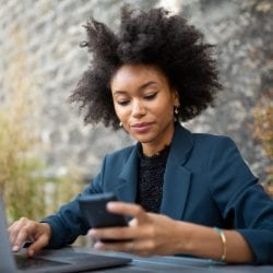 A professional woman sits in front of her laptop holding her cell phone.