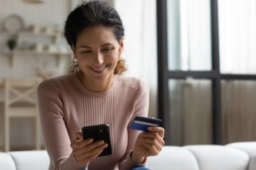 A young woman smiles down at her phone while holding a credit card.