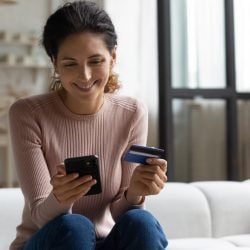 A young woman smiles down at her phone while holding a credit card.