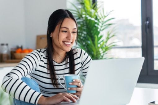 Happy young woman sitting at her table, focused on her laptop, and enjoying a cup of coffee