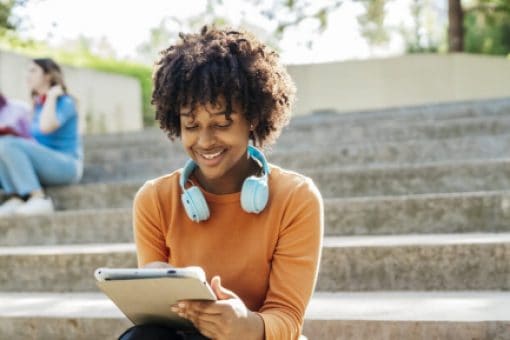 Woman with headphones around her neck, sitting on outdoor steps, engrossed in her tablet