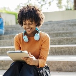 Woman with headphones around her neck, sitting on outdoor steps, engrossed in her tablet.