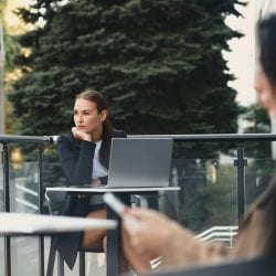 Woman sits outside at a table looking away from her laptop.