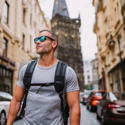 Man in sunglasses wearing backpack walks down European street.