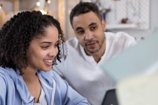 A young woman looks at a laptop with an older man in a brightly lit room.