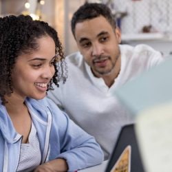 A young woman in a brightly lit room looks at a laptop with her father.