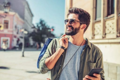 A smiling man walks down the street holding a shopping bag and a cell phone.