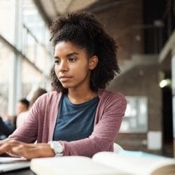 Young woman uses a laptop at a sunlit table with an open book beside her in a spacious office setting.