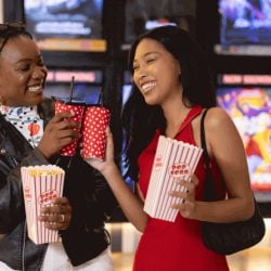 Two women with popcorn and drinks in a movie theater lobby.