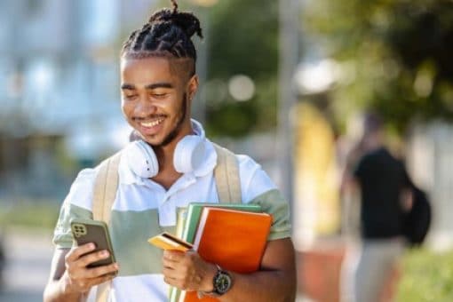 Smiling college student with dreadlocks using smartphone while holding textbooks and wearing headphones around neck on a sunny campus walkway.