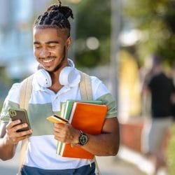 Smiling college student uses his smartphone on a sunny campus walkway.
