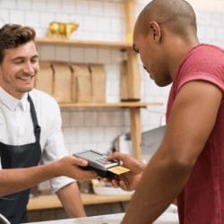 Barista handing a card reader to a customer making a payment with a credit card at a coffee shop counter.