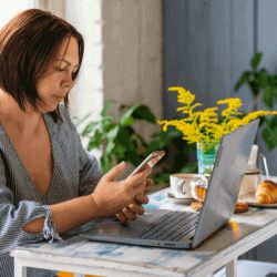 A woman sits at a table with a laptop and phone, and breakfast items and flowers on the table.
