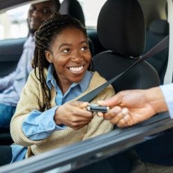 A person handing car keys to a couple who are sitting in a car looking happy.