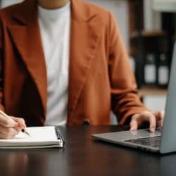 Person sits in front of laptop and writes in a notebook.