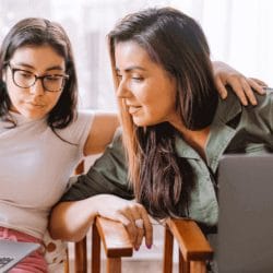 Two women sit together, looking at laptop screens, with one placing her arm around the other’s shoulders.