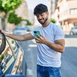 Smiling man staring at his phone leans on a car with one arm.