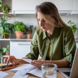 A woman uses a calculator on her mobile phone while holding a paper in her other hand. There's an open laptop on the table in front of her.