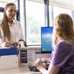 A woman stands at a hospital reception desk writing on paper.