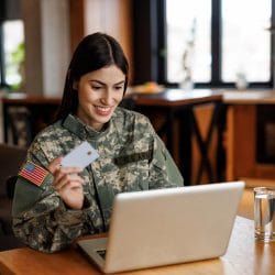 A woman in a military uniform sits at a table looking at a laptop while holding a credit card.